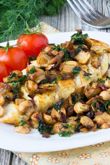 Garlic herb chicken breast topped with sautéed mushrooms, caramelized onions, and chopped green herbs on a white plate, with three vine tomatoes on the side. Rustic wood table background with fresh dill and a fork in the corner.