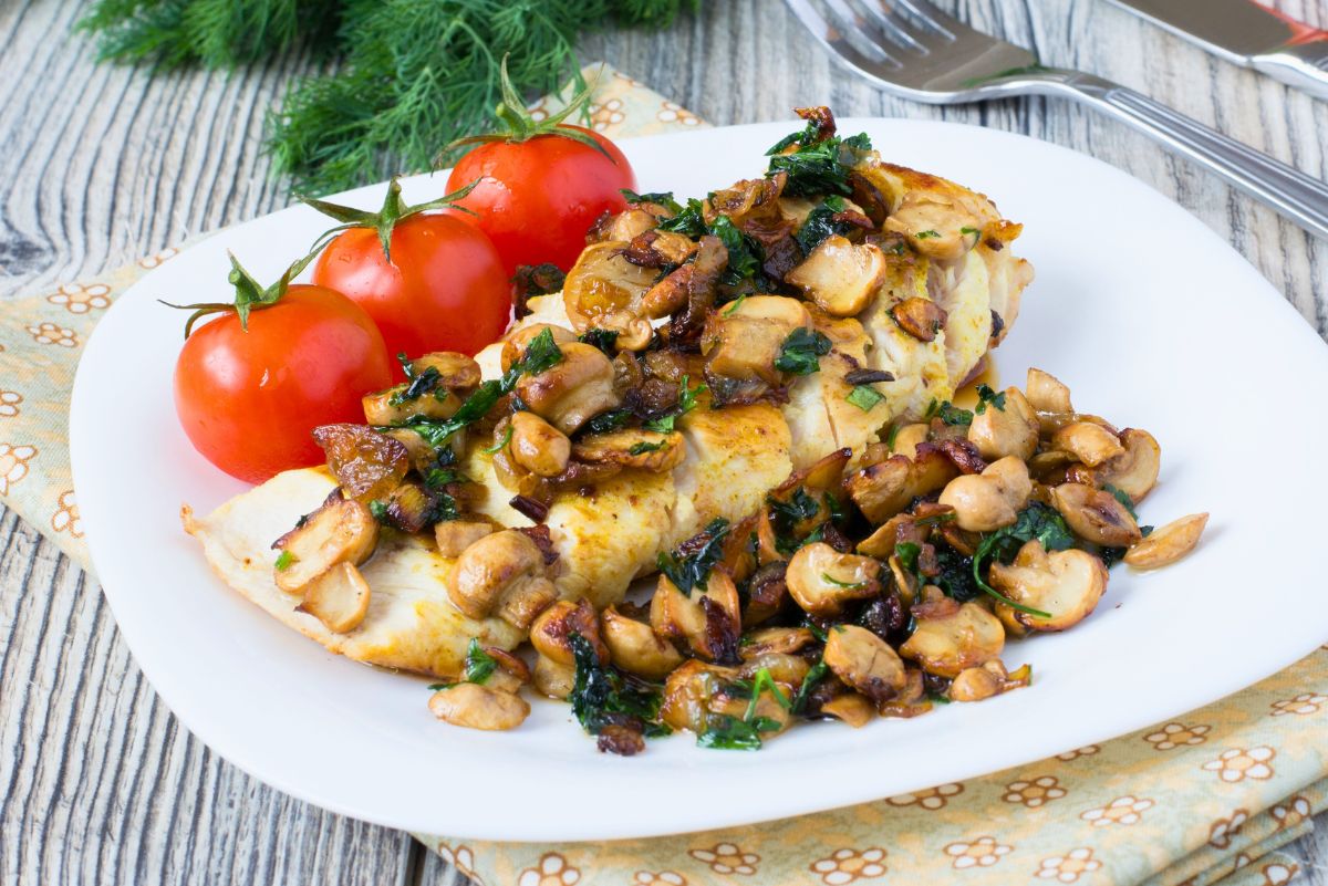 Garlic herb chicken breast topped with sautéed mushrooms, caramelized onions, and chopped herbs on a white plate, with three vine tomatoes on the side. Rustic wood table setting with a patterned cloth napkin, fresh dill in the background, and a fork near the top corner.