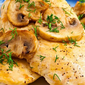 Close-up of garlic mushroom chicken skillet with juicy pan-seared chicken breasts topped with sautéed sliced mushrooms, fresh dill, cracked black pepper, and buttery garlic sauce on a blue-rimmed plate.