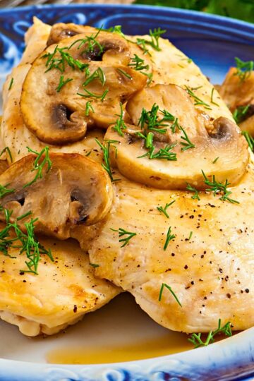 Close-up of garlic mushroom chicken skillet with juicy pan-seared chicken breasts topped with sautéed sliced mushrooms, fresh dill, cracked black pepper, and buttery garlic sauce on a blue-rimmed plate.
