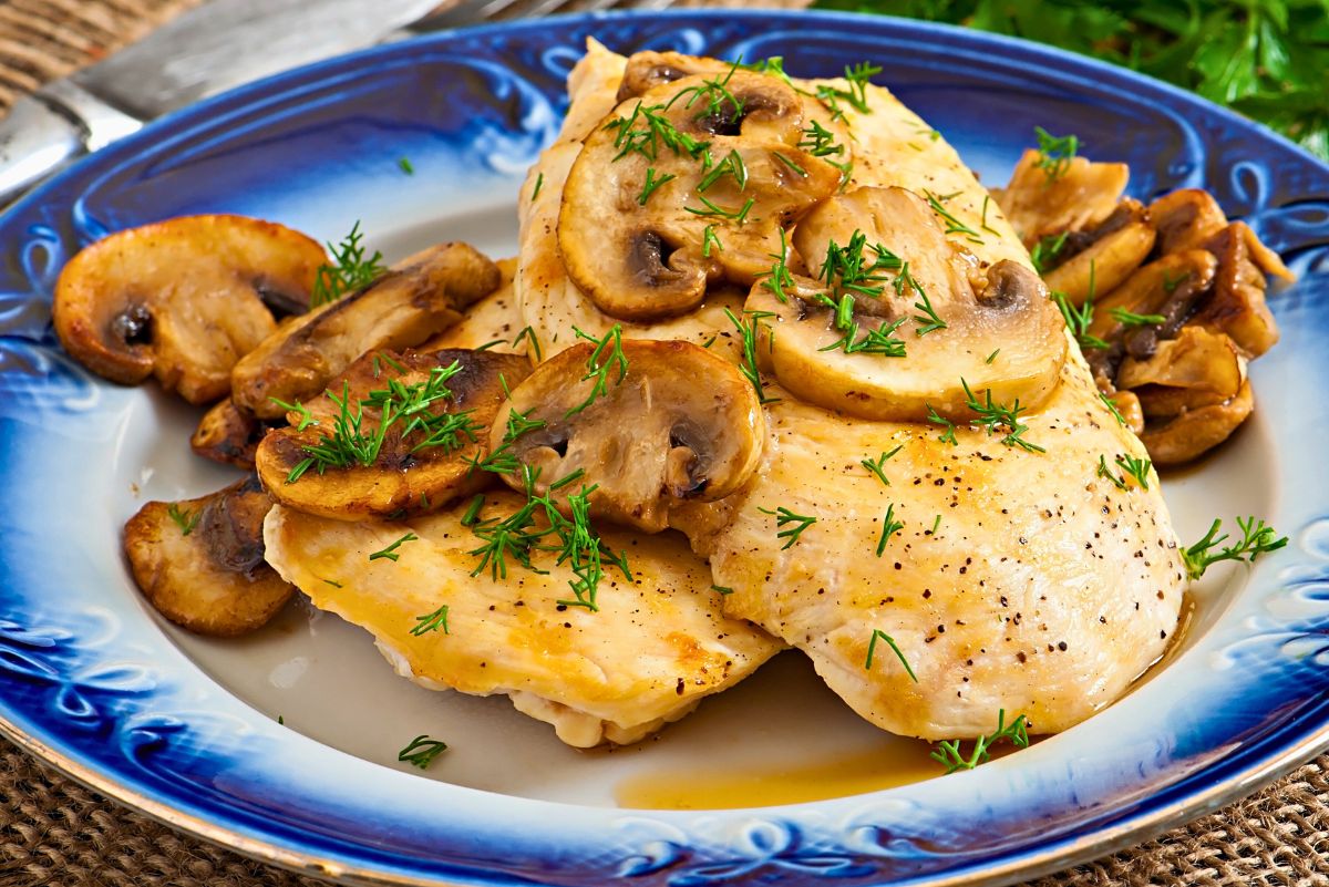 Garlic mushroom chicken skillet plated on a blue decorative dish with tender chicken breasts, sautéed mushroom slices, fresh dill, black pepper, and light garlic butter sauce. Rustic table setting with knife in the background.