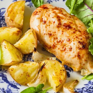 Garlic paprika baked chicken breast on a blue and white floral plate with roasted potato wedges, onion pieces, and fresh greens. Close-up dinner plate on a rustic wood table with golden roasted potatoes and juicy seasoned chicken.