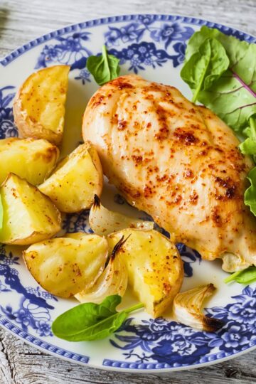 Garlic paprika baked chicken breast on a blue and white floral plate with roasted potato wedges, onion pieces, and fresh greens. Close-up dinner plate on a rustic wood table with golden roasted potatoes and juicy seasoned chicken.