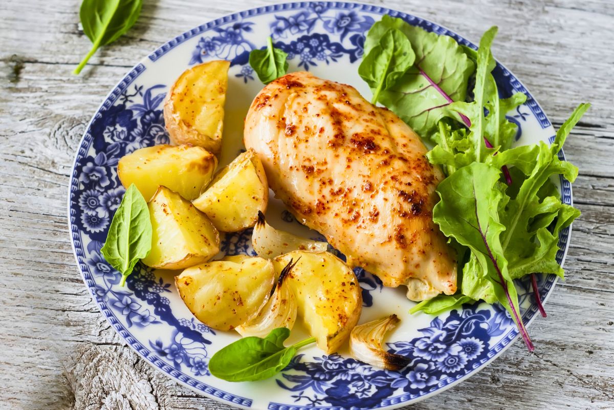 Garlic paprika baked chicken breast served with roasted potato wedges, onion, and mixed greens on a blue and white floral plate. Overhead dinner photo on a rustic wood table with juicy seasoned chicken and golden roasted potatoes.