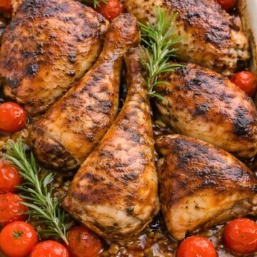 Garlic soy baked chicken thighs and drumsticks in a white baking dish, glazed with a dark glossy sauce and roasted until browned. Cherry tomatoes and fresh rosemary sprigs are tucked around the chicken, with pan juices and bits of garlic visible at the bottom.