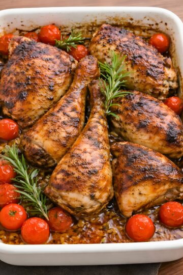 Garlic soy baked chicken thighs and drumsticks in a white baking dish, glazed with a dark glossy sauce and roasted until browned. Cherry tomatoes and fresh rosemary sprigs are tucked around the chicken, with pan juices and bits of garlic visible at the bottom.