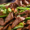 Close-up of high protein beef stir fry in a bowl, with chopsticks lifting a tender slice of beef over snow peas, carrots, and sliced green onions in a glossy sauce. Text on image: “nestinglane.com”.