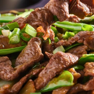 Close-up of high protein beef stir fry in a bowl, with chopsticks lifting a tender slice of beef over snow peas, carrots, and sliced green onions in a glossy sauce. Text on image: “nestinglane.com”.