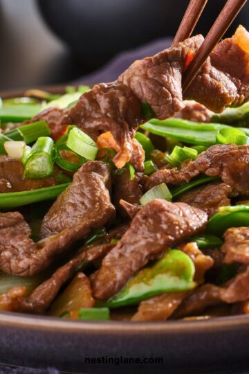 Close-up of high protein beef stir fry in a bowl, with chopsticks lifting a tender slice of beef over snow peas, carrots, and sliced green onions in a glossy sauce. Text on image: “nestinglane.com”.