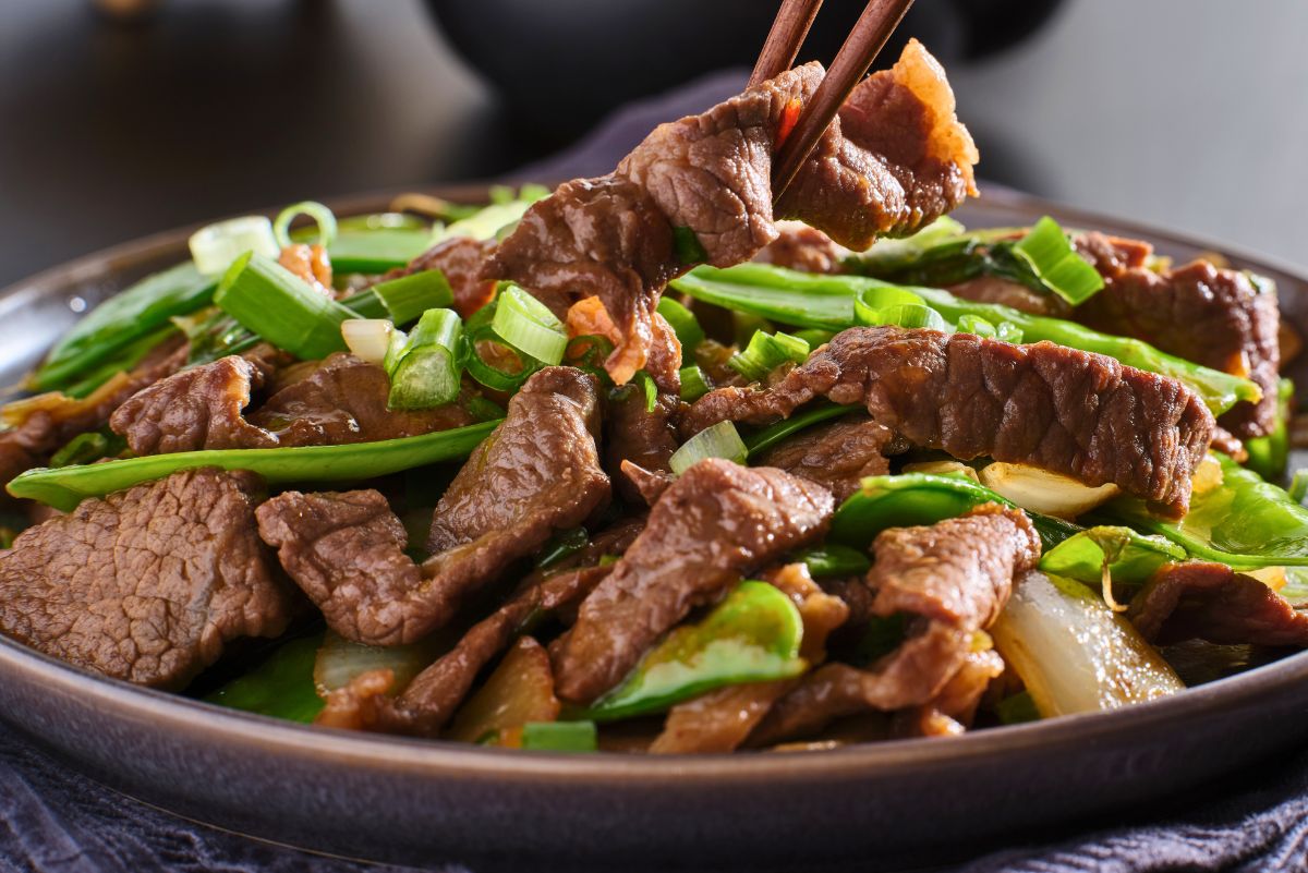 Bowl of high protein beef stir fry with sliced steak, snow peas, onions, and chopped green onions in a glossy sauce, with chopsticks lifting a piece of beef. No visible text. Quick beef stir fry recipe, healthy dinner idea.