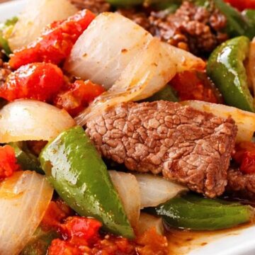 Close-up of crockpot pepper steak on a white plate with tender beef strips, chunky onion pieces, green bell peppers, and stewed tomatoes in a glossy sauce. High protein healthy dinner recipe photo on a rustic wood table.