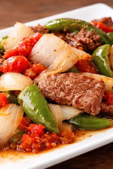 Close-up of crockpot pepper steak on a white plate with tender beef strips, chunky onion pieces, green bell peppers, and stewed tomatoes in a glossy sauce. High protein healthy dinner recipe photo on a rustic wood table.