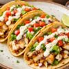 Three grilled fish tacos on corn tortillas, filled with flaky white fish, shredded lettuce, diced tomato, red onion, and creamy lime sauce, served on a rustic plate. Lime wedges, extra sauce, and pico de gallo sit in the background. High protein fish tacos with lime sauce food photo.