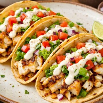 Three grilled fish tacos on corn tortillas, filled with flaky white fish, shredded lettuce, diced tomato, red onion, and creamy lime sauce, served on a rustic plate. Lime wedges, extra sauce, and pico de gallo sit in the background. High protein fish tacos with lime sauce food photo.
