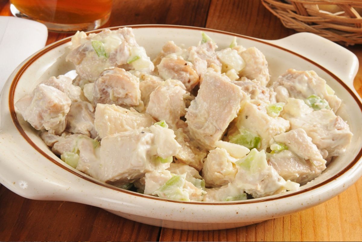 Chunky chicken salad in a white oval serving dish on a wooden table, with large bites of chicken and chopped celery in a creamy dressing. A glass of iced tea and a wicker basket sit blurred in the background.
