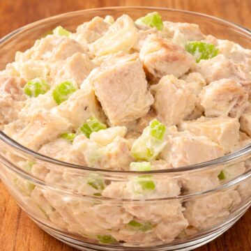 Chunky chicken salad in a clear glass bowl on a warm wooden table, with large pieces of chicken in a creamy dressing and small chopped celery mixed throughout. Close-up food photo with a simple homemade deli salad look.