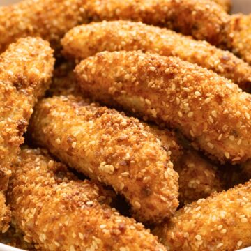 Close-up of crispy air fryer sesame chicken strips piled in a white bowl on a dark wood table. Golden breaded chicken tenders are coated with sesame seeds and shown in warm natural light.