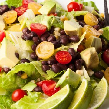Southwest black bean and avocado salad with romaine lettuce, black beans, cherry tomatoes, corn, avocado chunks, and lime wedges on a white plate. Fresh healthy lunch or vegetarian salad with bright green, red, and yellow ingredients.