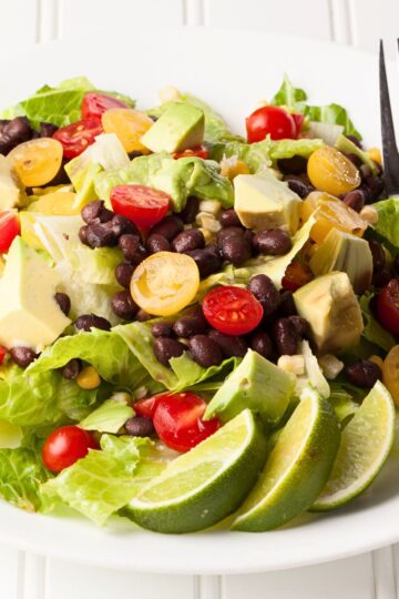 Southwest black bean and avocado salad with romaine lettuce, black beans, cherry tomatoes, corn, avocado chunks, and lime wedges on a white plate. Fresh healthy lunch or vegetarian salad with bright green, red, and yellow ingredients.