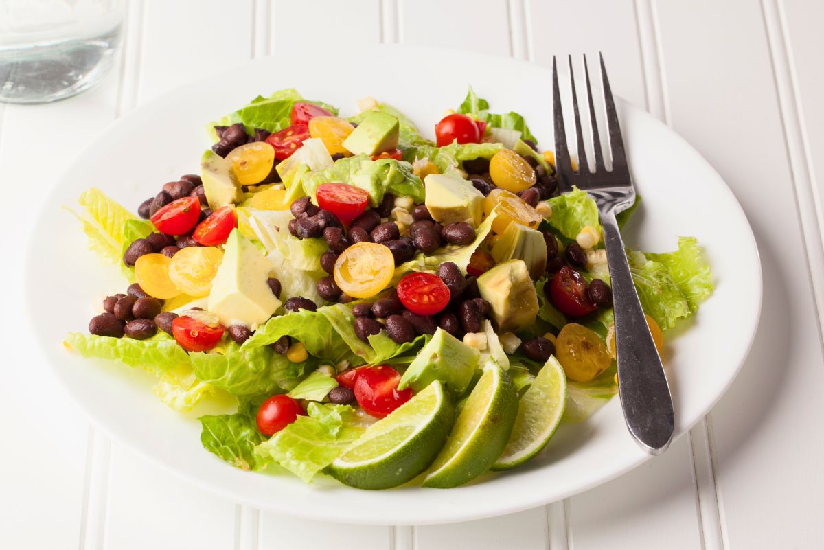 Southwest black bean and avocado salad on a white plate with romaine lettuce, halved cherry tomatoes, corn, avocado chunks, black beans, and lime wedges. A fork rests on the plate on a white tabletop.