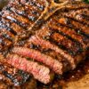 Close-up of grilled T-bone steak with a glossy sweet chili glaze on a wooden cutting board. Several slices show a juicy pink center, with charred edges and sauce pooling around the meat. A lemon wedge and leafy greens sit blurred in the background.