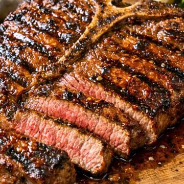 Close-up of grilled T-bone steak with a glossy sweet chili glaze on a wooden cutting board. Several slices show a juicy pink center, with charred edges and sauce pooling around the meat. A lemon wedge and leafy greens sit blurred in the background.