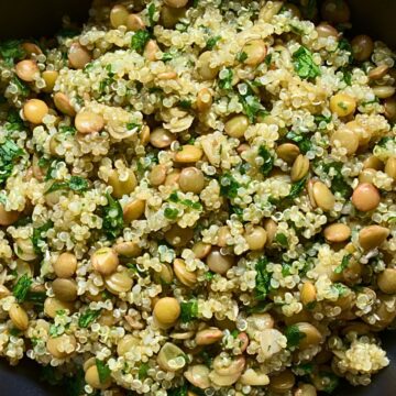 Overhead view of quinoa lentil salad in a black skillet, with fluffy quinoa, green lentils, and chopped fresh herbs mixed throughout. The healthy grain salad sits on a wood table with a spoon handle visible at the edge.