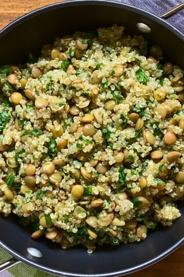 Overhead view of quinoa lentil salad in a black skillet, with fluffy quinoa, green lentils, and chopped fresh herbs mixed throughout. The healthy grain salad sits on a wood table with a spoon handle visible at the edge.