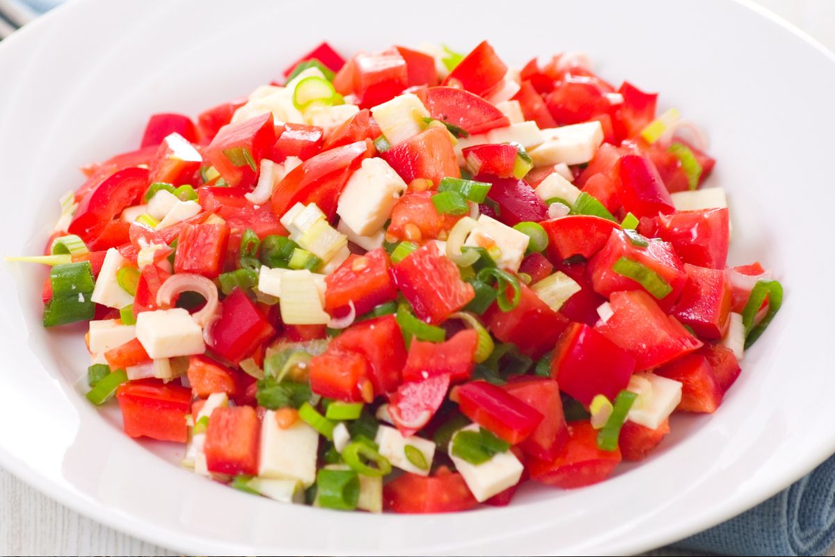 Fresh tomato and red pepper salad with mozzarella cubes and sliced green onion in a white bowl. Bright red chopped vegetables and creamy cheese make a simple summer salad on a light tabletop.