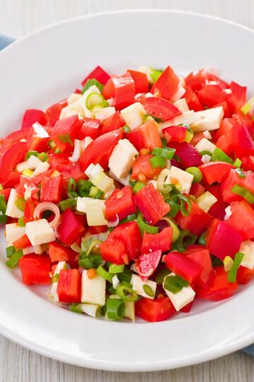 Tomato and red pepper salad in a white bowl, made with chopped tomatoes, red bell pepper, mozzarella cubes, and sliced green onion. Bright fresh salad close-up on a light wood surface, with juicy red pieces and green onion scattered throughout.