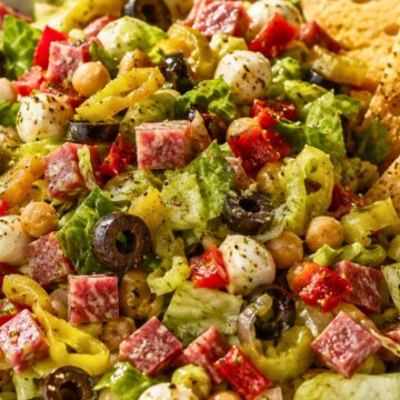 Antipasto chopped salad with romaine lettuce, salami, mozzarella pearls, chickpeas, black olives, banana peppers, red peppers, Italian dressing, and garlic toast chips in a white bowl.