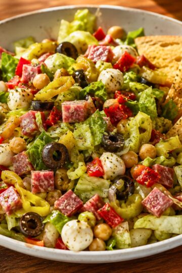 Antipasto chopped salad with romaine lettuce, salami, mozzarella pearls, chickpeas, black olives, banana peppers, red peppers, Italian dressing, and garlic toast chips in a white bowl.