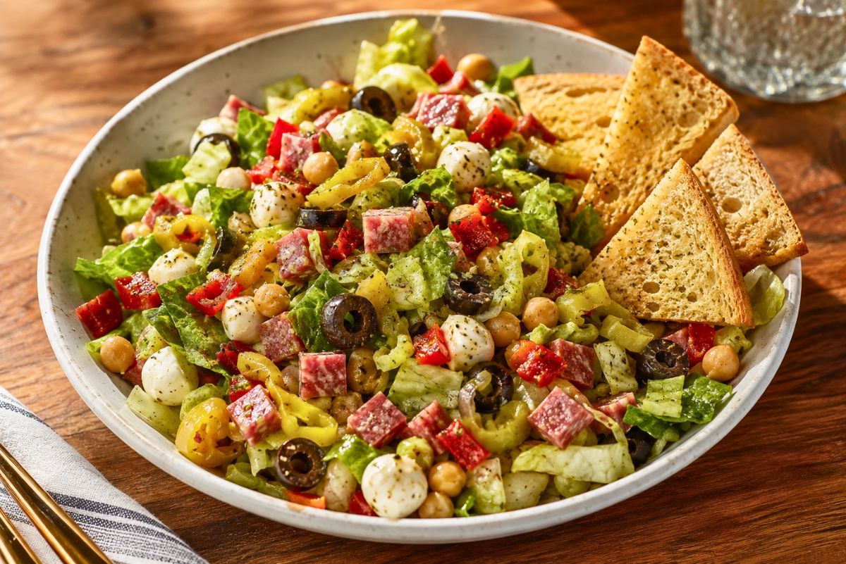 Antipasto chopped salad with romaine, salami, mozzarella pearls, chickpeas, black olives, banana peppers, red peppers, and Italian dressing, served in a white bowl with garlic toast chips on the side.
