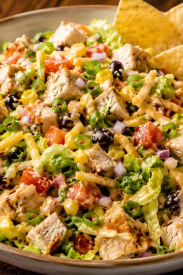 BBQ chicken chopped salad with grilled chicken, lettuce, black beans, corn, tomatoes, red onion, shredded cheese, green onions, and tortilla chips in a bowl.