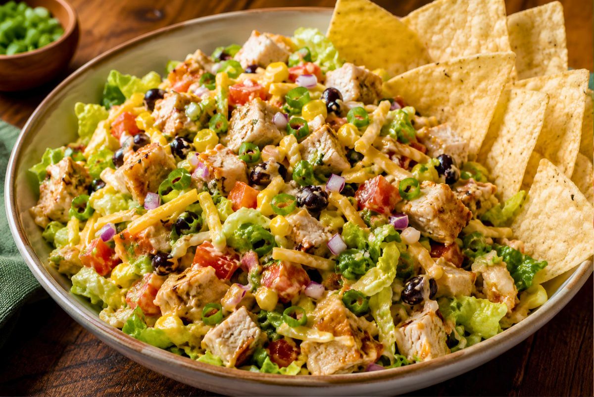 BBQ chicken chopped salad with grilled chicken, romaine lettuce, black beans, corn, tomatoes, red onion, shredded cheese, green onions, and tortilla chips served in a large bowl.