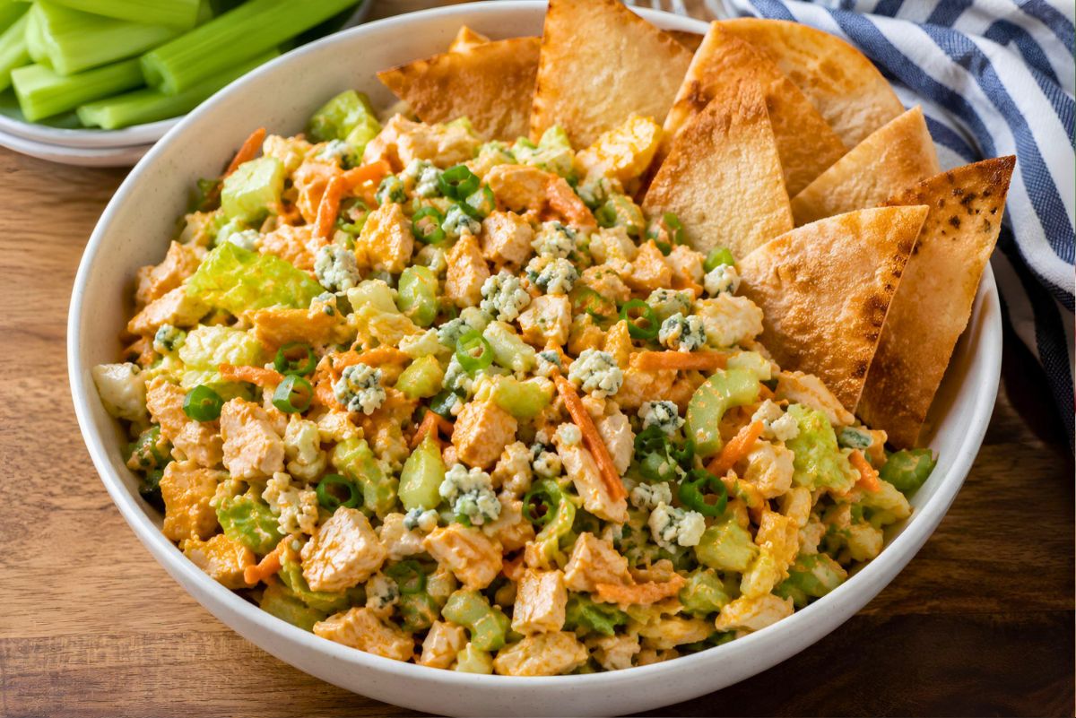 Buffalo chicken chopped salad in a white bowl with chopped chicken, lettuce, celery, carrots, green onions, blue cheese crumbles, buffalo ranch dressing, and toasted pita chips. A small bowl of celery sticks sits in the background.