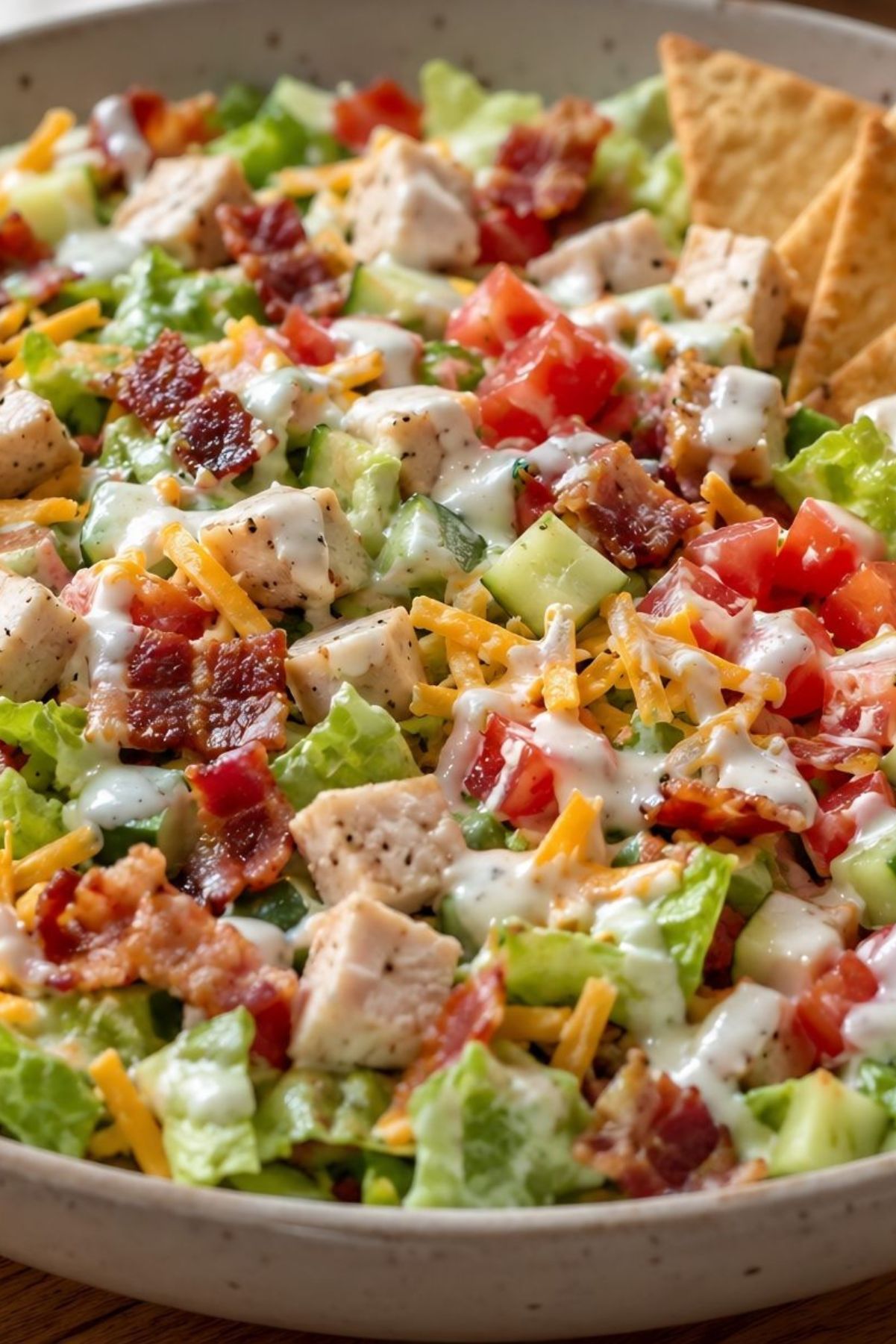 Chicken bacon ranch chopped salad with diced chicken, crisp bacon, lettuce, cucumbers, tomatoes, shredded cheddar cheese, creamy ranch dressing, and pita chips served in a bowl.