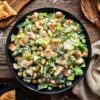 Chicken Caesar chopped salad with romaine, grilled chicken, chickpeas, Parmesan, and creamy Caesar dressing in a black bowl. Pita wedges, grated cheese, and a linen napkin surround the bowl on a rustic wooden table.