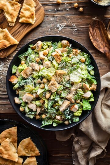 Chicken Caesar chopped salad with romaine, grilled chicken, chickpeas, Parmesan, and creamy Caesar dressing in a black bowl. Pita wedges, grated cheese, and a linen napkin surround the bowl on a rustic wooden table.