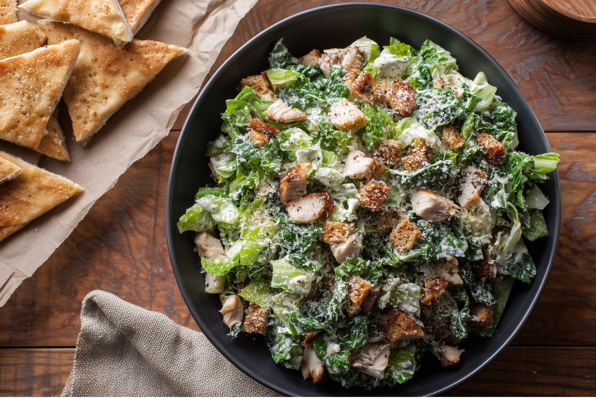 Chicken Caesar chopped salad in a black bowl with chopped romaine, grilled chicken, croutons, Parmesan cheese, and creamy Caesar dressing. Pita wedges sit on parchment beside the salad on a rustic wood table.