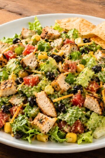 Chicken fajita chopped salad with grilled chicken, romaine lettuce, black beans, corn, tomatoes, cheddar cheese, creamy dressing, and tortilla chips on a white plate.