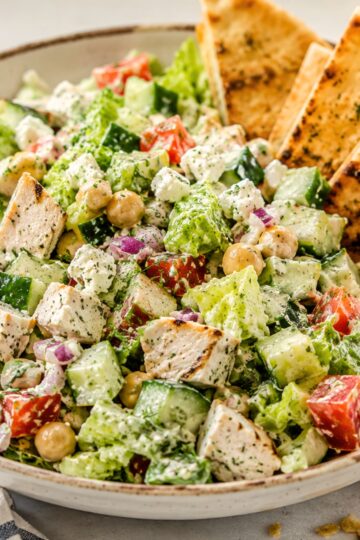 Chicken gyro chopped salad with grilled chicken, romaine, cucumber, tomatoes, chickpeas, red onion, feta, creamy tzatziki-style dressing, and toasted pita chips in a shallow bowl.