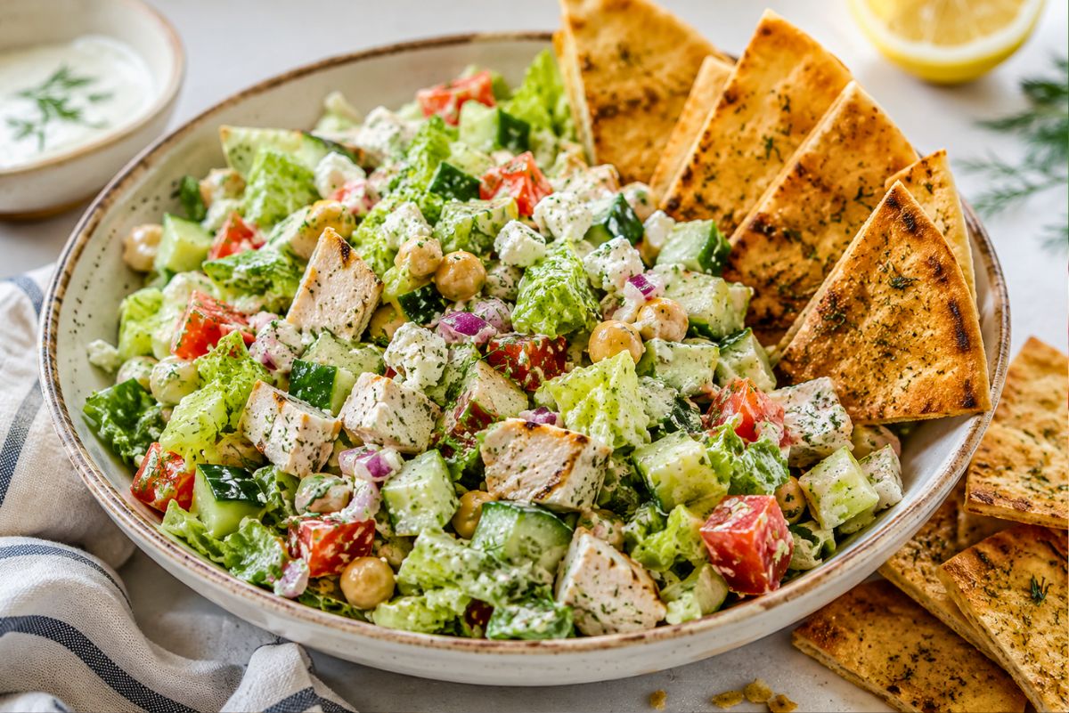 Chicken gyro chopped salad with grilled chicken, romaine, cucumbers, tomatoes, chickpeas, red onion, feta, creamy tzatziki-style dressing, and toasted pita chips in a wide bowl with lemon and dip nearby.