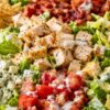Cobb chopped salad with grilled chicken, crispy bacon, diced tomatoes, blue cheese crumbles, romaine lettuce, creamy dressing, and toasted pita chips on the side in a white bowl.