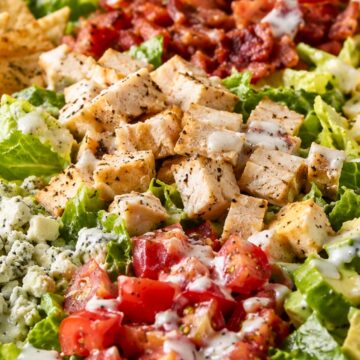 Cobb chopped salad with grilled chicken, crispy bacon, diced tomatoes, blue cheese crumbles, romaine lettuce, creamy dressing, and toasted pita chips on the side in a white bowl.