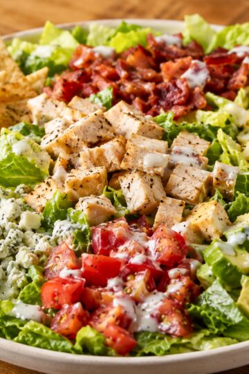 Cobb chopped salad with grilled chicken, crispy bacon, diced tomatoes, blue cheese crumbles, romaine lettuce, creamy dressing, and toasted pita chips on the side in a white bowl.