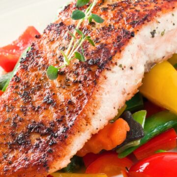 Crispy pan-seared salmon fillet with a golden herb crust served over colorful garden vegetables, including bell peppers, carrots, green beans, peas, and fresh herbs.