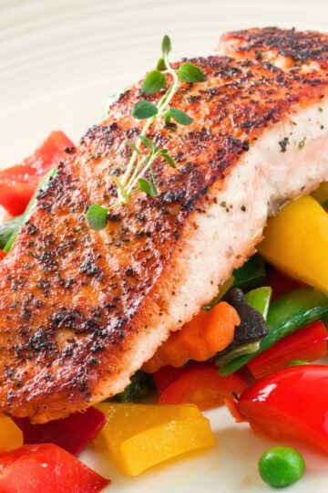 Crispy pan-seared salmon fillet with a golden herb crust served over colorful garden vegetables, including bell peppers, carrots, green beans, peas, and fresh herbs.