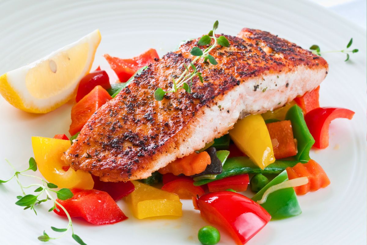 Crispy pan-seared salmon with a golden herb crust served over colorful garden vegetables, red and yellow bell peppers, green beans, carrots, peas, and a lemon wedge.