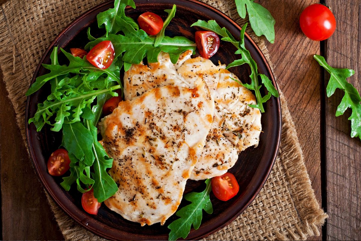 Garlic herb chicken breast on a dark plate with fresh arugula and cherry tomatoes, served on a rustic wooden table for a healthy high protein chicken salad dinner.